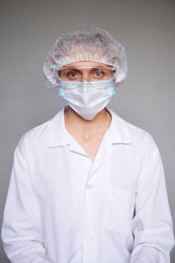 Tıbbi maskeli, cerrahi şapkalı ve gri arka planda gözlüklü kadın doktor ya da bilim adamına yakın durun..