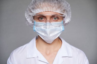 Tıbbi maskeli, cerrahi şapkalı ve gri arka planda gözlüklü kadın doktor ya da bilim adamına yakın durun..