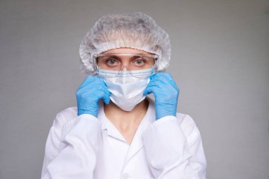 Tıbbi maskeli, cerrahi şapkalı, gözlüklü ve eldivenli kadın doktor ve bilim adamlarına yakın plan gri arka plan.
