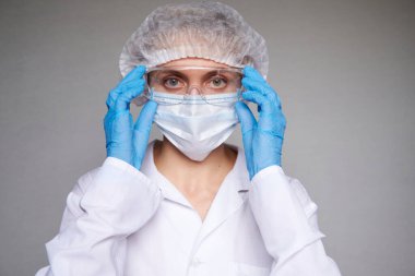 Tıbbi maskeli, cerrahi şapkalı, gözlüklü ve eldivenli kadın doktor ve bilim adamlarına yakın plan gri arka plan.
