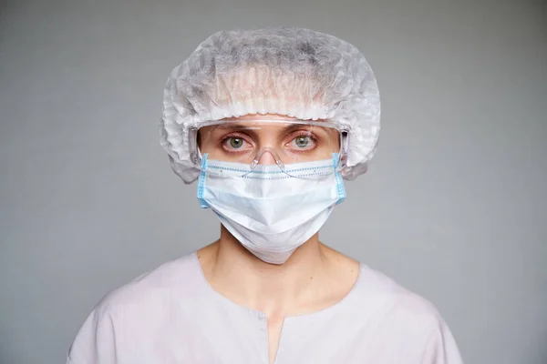 Tıbbi maskeli, cerrahi şapkalı ve gri arka planda gözlüklü kadın doktor ya da bilim adamına yakın durun..