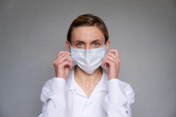 Close up of female doctor or scientist in protective facial mask over grey background. She is adjusting the medical mask with her hands