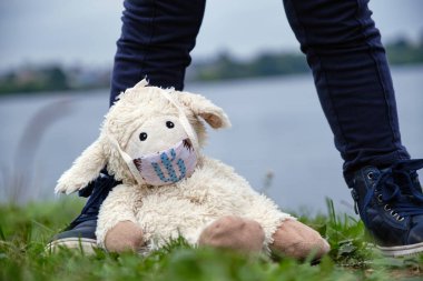 Maskeli küçük kız maske takmış oyuncağıyla gölün kenarında duruyor.