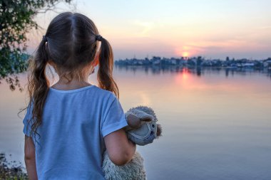 Maskeli küçük kız, gün batımının arkasındaki maske takmış oyuncağıyla gölün kenarında duruyor.