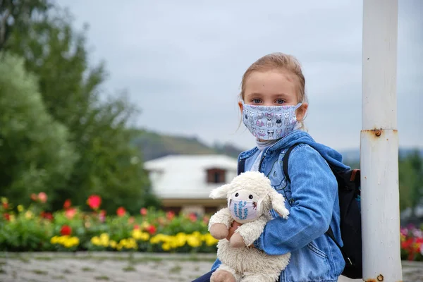 Maskeli küçük bir kız lamba direğinin yanında oturuyor ve elinde maske takan bir oyuncak tutuyor.