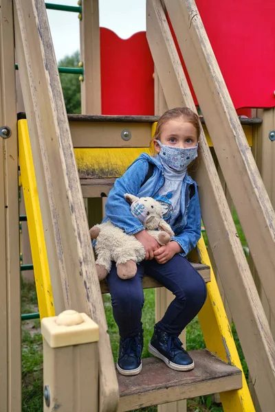Maskeli küçük kız maske takmış oyuncağıyla parkta oturuyor.