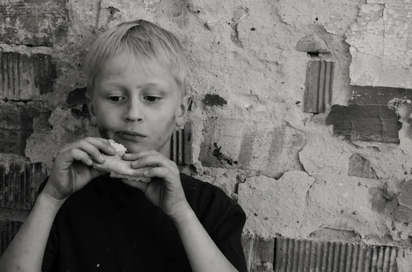 Aç bir kirli çocuk bir kabuk ekmek duvara iştahla yiyor. Siyah ve beyaz.