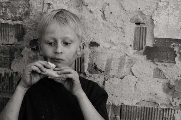 Голодный грязный мальчик жадно съедает корку хлеба у стены. Черное и белое
.