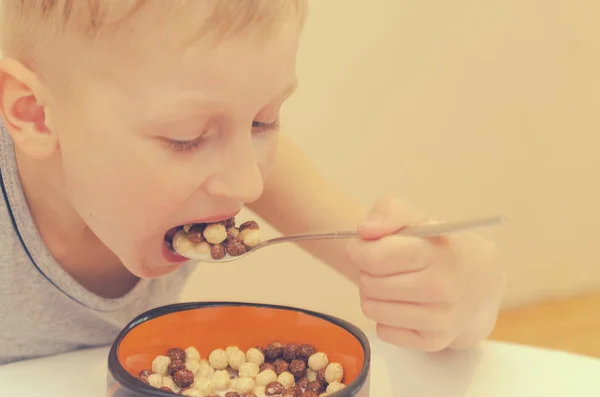 Un niño con una camiseta gris desayuna copos de maíz con leche . — Foto de Stock