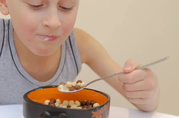 Un niño con una camiseta gris desayuna copos de maíz con leche . — Foto de Stock