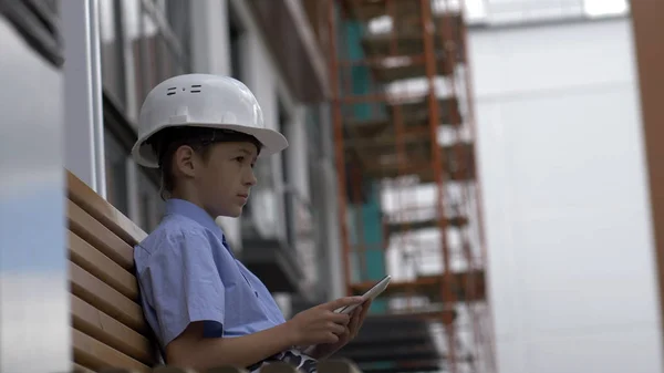 boy builder sits on the bench uses a tablet, monitors the builder project, plays a builder