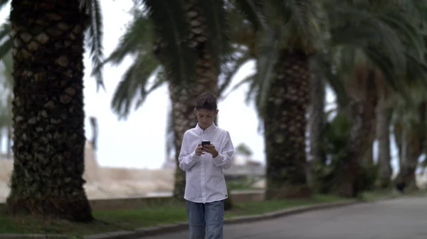 boy goes on the road past the palm trees and uses the phone, is chatting with someone on the phone