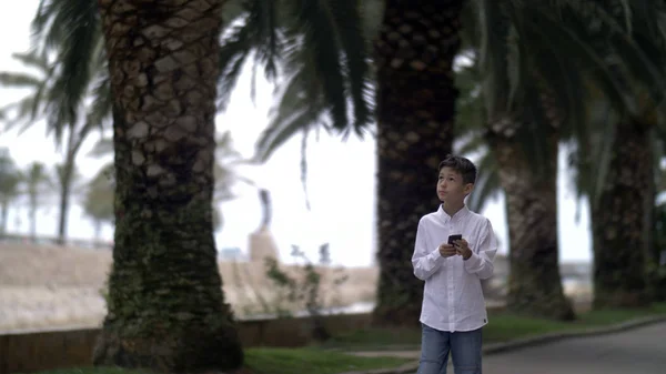 boy goes on the road past the palm trees and uses the phone, is chatting with someone on the phone