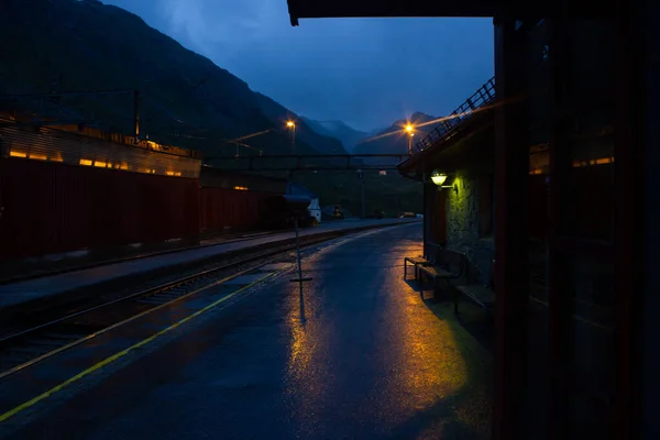 Boş tren istasyonunun caddesinde bulutlu bir akşam. Tamamen yalnız. Demiryolları ve dağlar görünür..