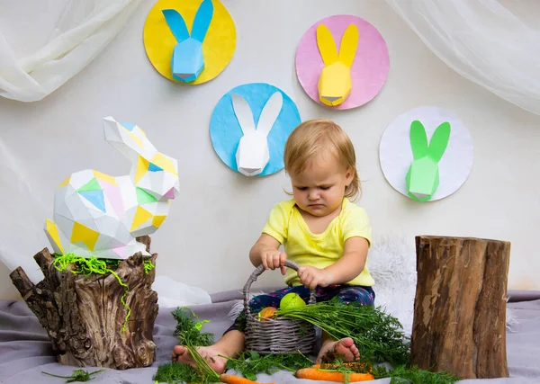 Sarı tişörtlü küçük bir çocuk havuçla oynar Paskalya dekorunun arka planına karşı, paskalya tavşanları kağıttan yapılır.