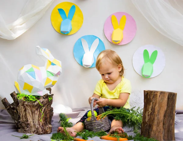 Sarı tişörtlü küçük bir çocuk havuçla oynar Paskalya dekorunun arka planına karşı, paskalya tavşanları kağıttan yapılır.