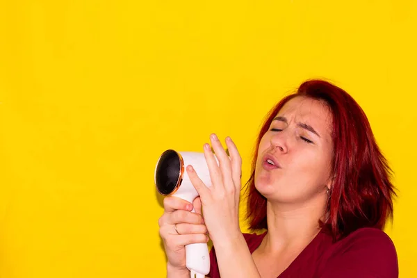 Sarı arka planda kızıl saçlı kız saç kurutma makinesinde şarkı söylüyor.