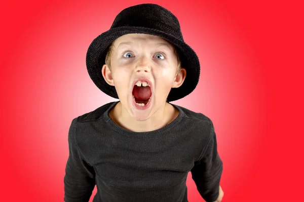 黑色衬衫和黑色帽子的小男孩特写镜头 沉重的尖叫表情和眼睛睁得大大的 疯狂的看红色背景 — 图库照片