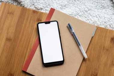 Ahşap bir tahta üzerinde bir defter ve kalem ile boş ekran akıllı telefonunun ayrıntıları. Mobil ekran boş.