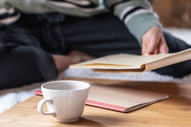 Evde defterlerle çalışan ve yerde sıcak bir içecekle oturan bir kız..