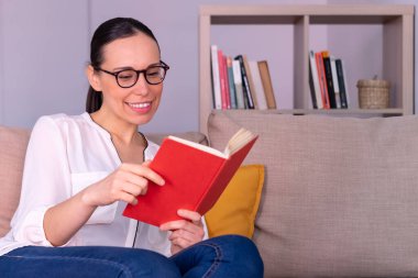 Evdeki kanepede oturmuş kitap okurken gülümseyen güzel esmer kadın. Beyaz bir gömlek, gözlük ve at kuyruğu takıyor..