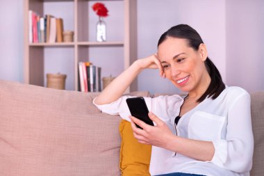 Evdeki kanepede oturan akıllı telefona gülümseyen güzel esmer kadın. Beyaz bir gömlek ve at kuyruğu giyiyor..