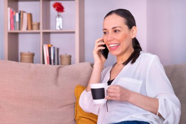 Evdeki kanepede oturmuş akıllı telefondan konuşan mutlu, güzel, esmer kadın..