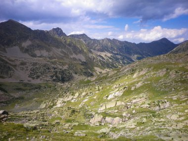 Gün boyunca yüksek bir dağ vadisinin manzarası. Ağaçlarla dolu. Uzakta dağlar ve gökte bulutlar var. Yatay resim