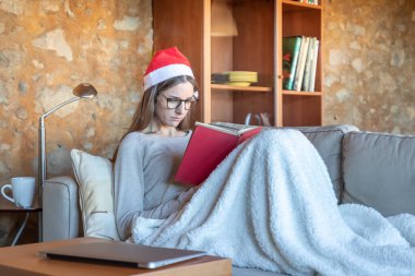 Evdeki kanepede oturan üzgün, güzel, esmer bir kadın Noel Baba şapkasıyla battaniyeyle kaplı bir kitap okuyor. Gözlük takıyor..