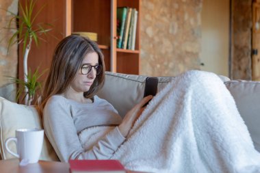 Üzgün, genç, güzel, esmer kadın evdeki kanepede battaniyeyle oturan akıllı telefona bakıyor. Gözlük takıyor..