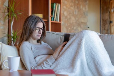 Evdeki kanepede battaniyeyle oturmuş akıllı telefona bakan genç ve güzel bir esmer kadın. Gözlük takıyor..