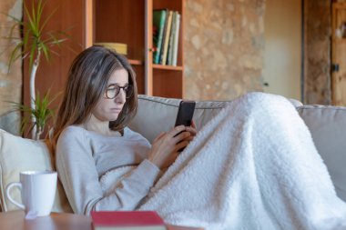 Evdeki kanepede battaniyeyle oturan akıllı telefona bakan hayal kırıklığına uğramış genç, güzel esmer kadın. Gözlük takıyor..