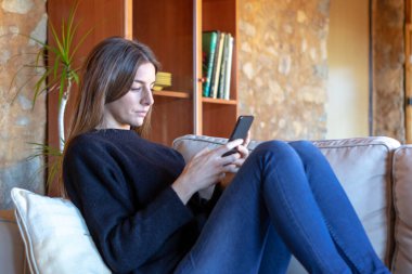 Ciddi esmer, genç bir kadın. Evdeki kanepede oturmuş akıllı telefonla sohbet ediyor. Siyah kazak ve kot pantolon giyiyor.