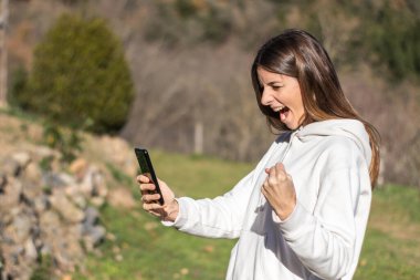 Güneşli bir günde güzel bir esmer kadın parktaki akıllı telefondan haber okuyor. Beyaz bir kazak giyiyor ve uzun saçları var..