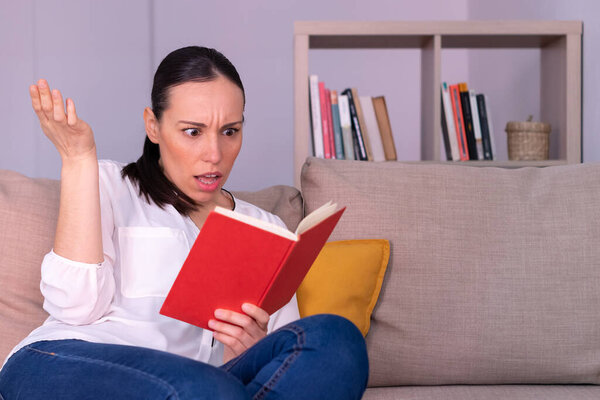 Pretty young brunette woman confused reading a book sitting on the sofa at home. She is wearing a white shirt and a ponytail.