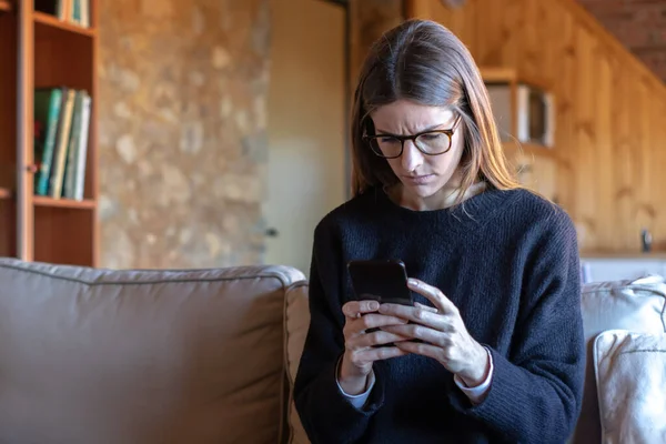 Confused young pretty brunette woman with glasses typing on smartphone sitting on sofa at home wearing black sweater
