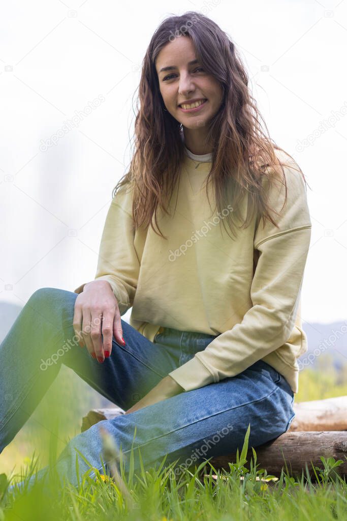 Pretty young brunette woman smiling and looking at camera sitting in a park outdoors on a sunny day. She is wearing blue jeans and a yellow sweater