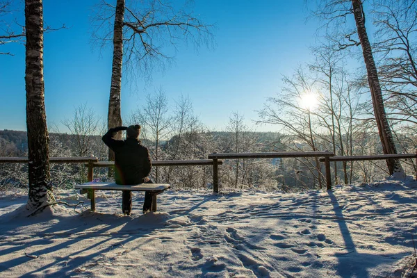 man welcoming fosun rising in heavy snow covered foirst light, sitting on wooden bench. est. first rays of light shining on frost iced tree branches and coloring vast landscape