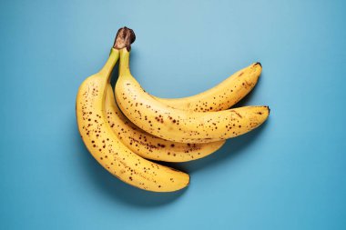 Kahverengi benekli üç sarı muzdan oluşan bir küme parlak mavi bir yüzeye dayanır. Olgunluklarını ve tüketimi davet eden çekici dokularını sergiler..