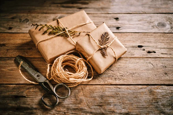 Kahverengi kâğıt ve sicimle sarılı iki güzel hediye kırsal bir ahşap masaya yerleştirilir. Makas ve doğal süslemeler sahneye cazibe katar..