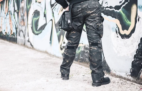 An urban man in a camouflage military pants stands next to the wall. Background from the urban part of the city