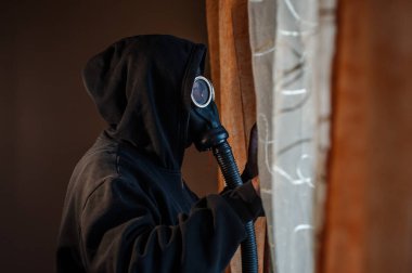 Siyah kapüşonlu ve koruyucu gaz maskeli kimliği belirsiz bir adam pencerenin önünde dikilip pencere kenarına bakıyor.