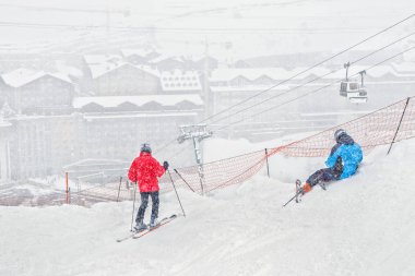 İki kayakçı bir yamaçta yoğun kar yağışı sırasında dinlen. Bir yamaç teleferik hattı ve kayak merkezi bina görünür