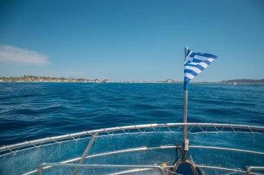 Flag of Greece waving over the see on cruise sheep in front of beautiful coastline on a sunny day.