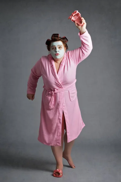 funny overweight fifty year old woman wearing bathrobe and face mask chasing a mosquito with a slipper on studio grey background