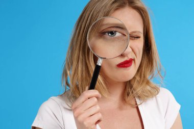 Genç ve beyaz tenli sarışın kadın gülümsüyor ve mavi izole stüdyo arka planındaki büyüteçten bakıyor.