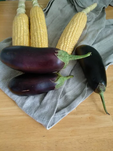 成熟的紫色茄子和没有叶子的黄色玉米躺在木制桌子上的灰色亚麻布毛巾上 — 图库照片