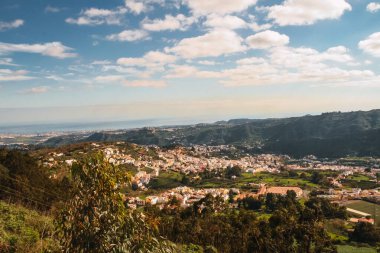 Büyük Kanarya Adasının dağlarındaki küçük bir köyün manzarası. Kayalar, çalılar, farklı türde çiçekler, ağaçlar ve yoğun mavi renkte bulutlu gökyüzü.