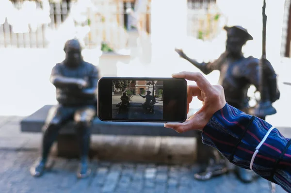 Woman holding cell phone with the picture of Don Quixote and Sancho Panza metal sculptures settled one in front of the other in main street of Alcala de Henares