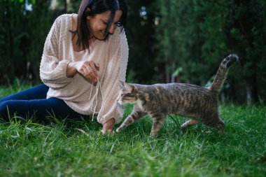 Kadın çimenlerde oturup kedi ile oynarken gülümsüyor ve ip sunuyor. Seçici Odaklanma. Ev hayatı işte. Hayvan sevgisi